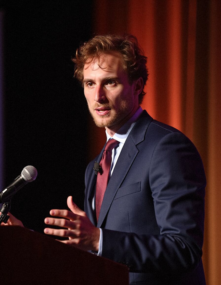 AI headshot of Theo: at a lectern, navy suit and red tie